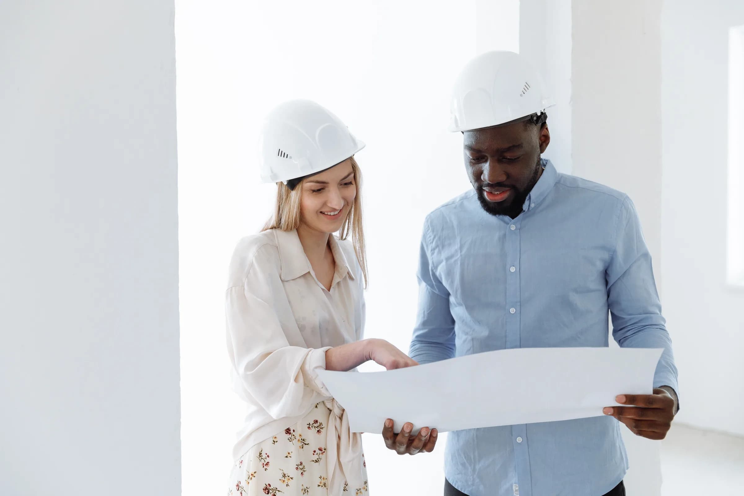 Two engineers in hard hats read a plan in a bright, unfinished room.