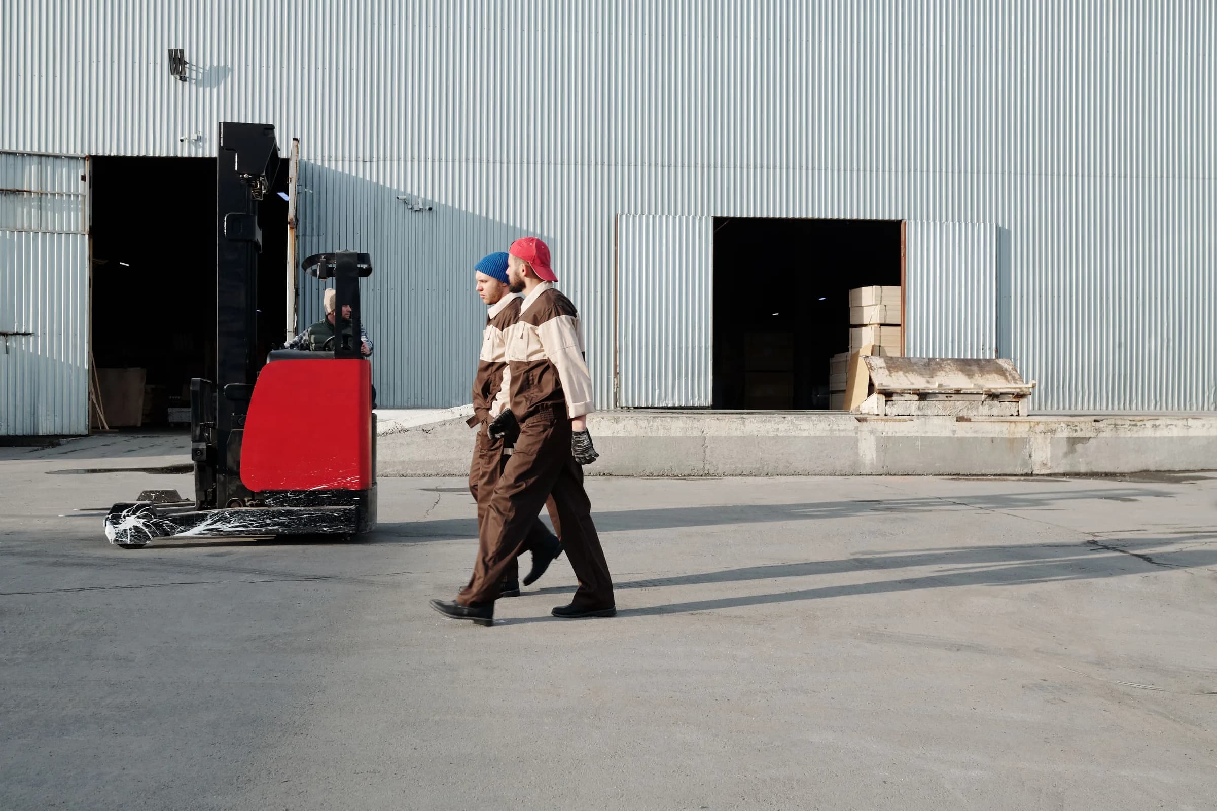 Two warehouse workers cross a sunlit yard beside a forklift.